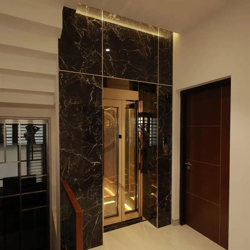Black marble clad residential foyer with open glass elevator doors revealing a mirrored, gold lit cabin interior next to a wooden entry door.