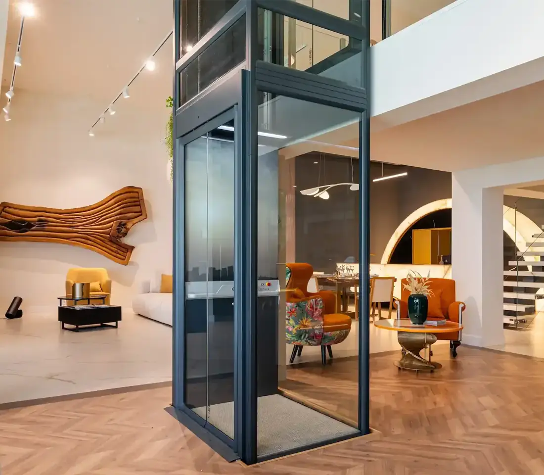 Modern foyer elevator with dark stone clad walls, glass cabin doors open beside a glass railed staircase.