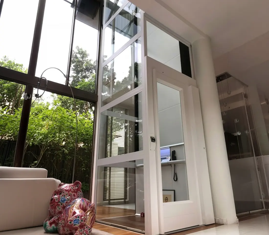 Modern foyer elevator with dark stone clad walls, glass cabin doors open beside a glass railed staircase.