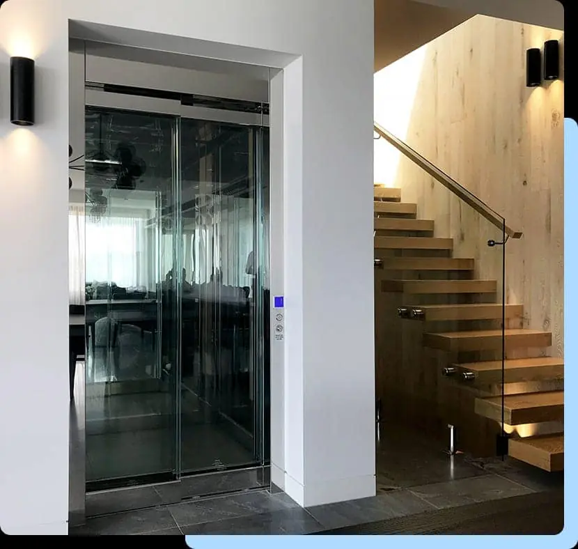 Glass walled ESTELLA cabin set into a wall alcove beside an open tread wooden staircase with a wall mounted control panel.