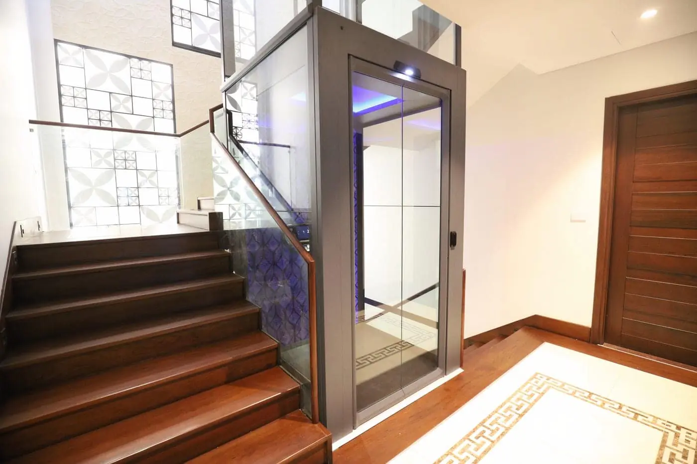 Grey framed glass ESTELLA cabin with blue interior lighting beside a wooden staircase, opening onto a marble inlay entrance floor.