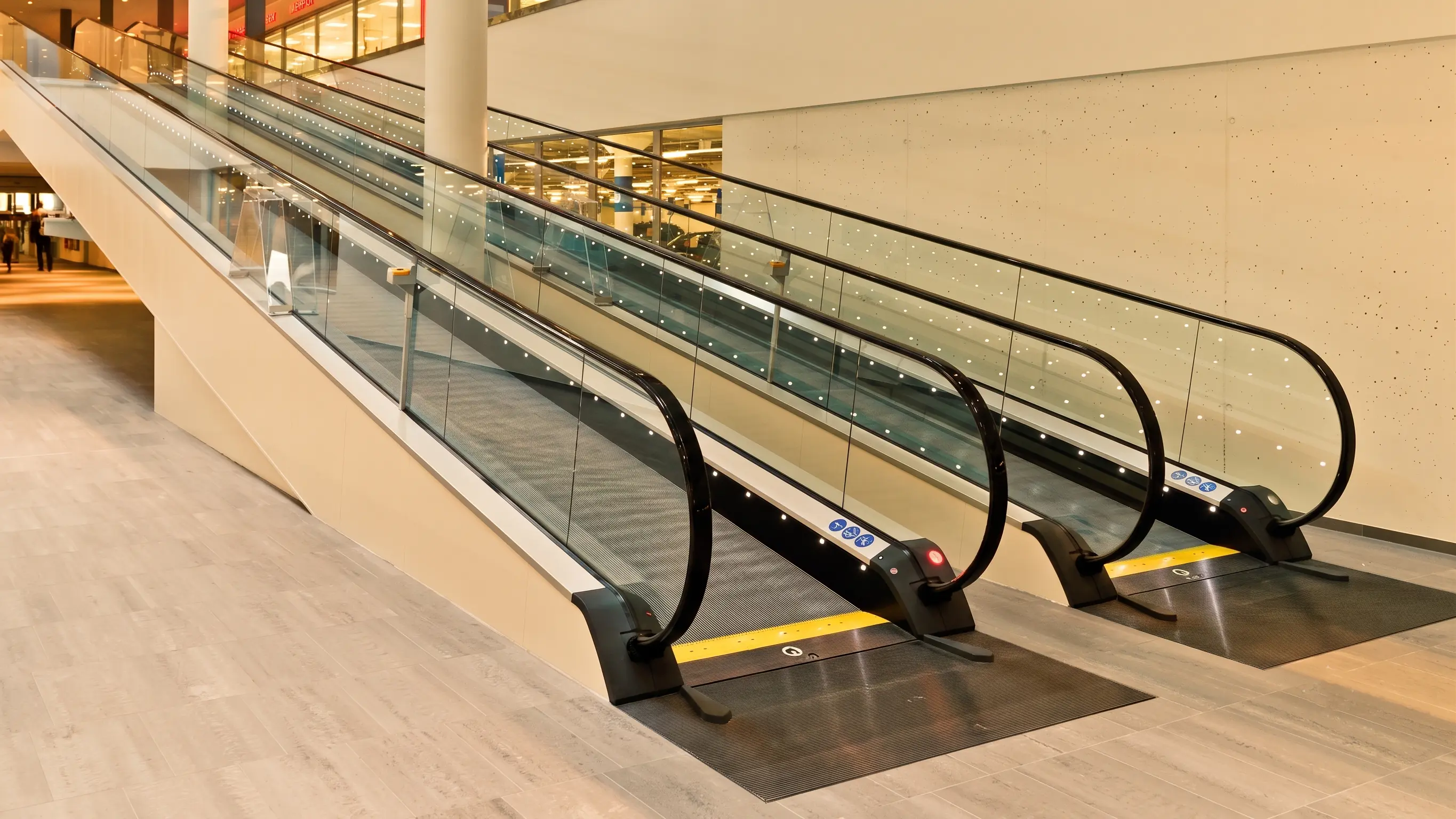 Twin inclined travelators rising through an empty mall concourse toward glass balustrades and storefronts.