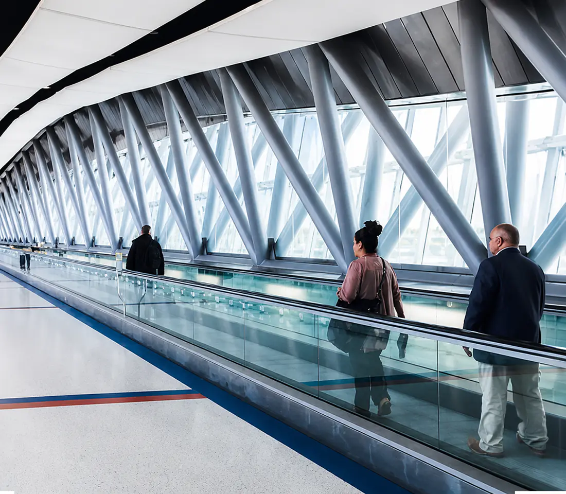 Passengers ride a curved moving walkway beneath a triangulated glass and steel terminal roof.