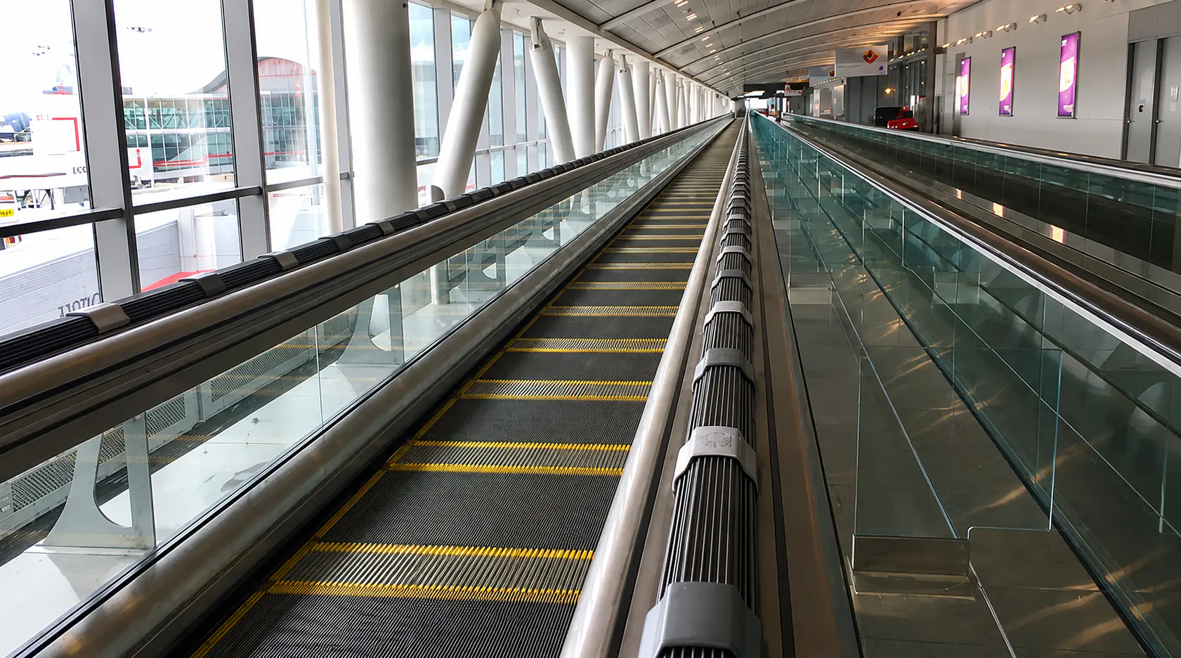 Twin inclined travelators rising through an empty mall concourse toward glass balustrades and storefronts.
