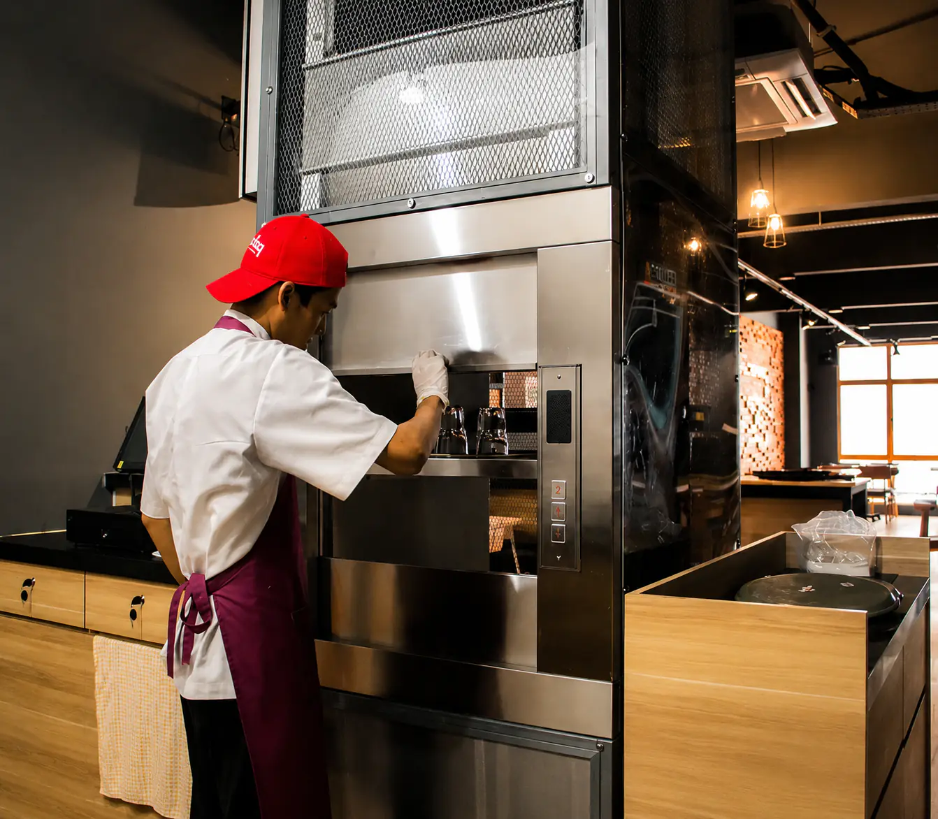 Stainless steel service lift standing open in a restaurant kitchen, its shelf loaded with a chafing dish and stacked plates beside the prep stations.