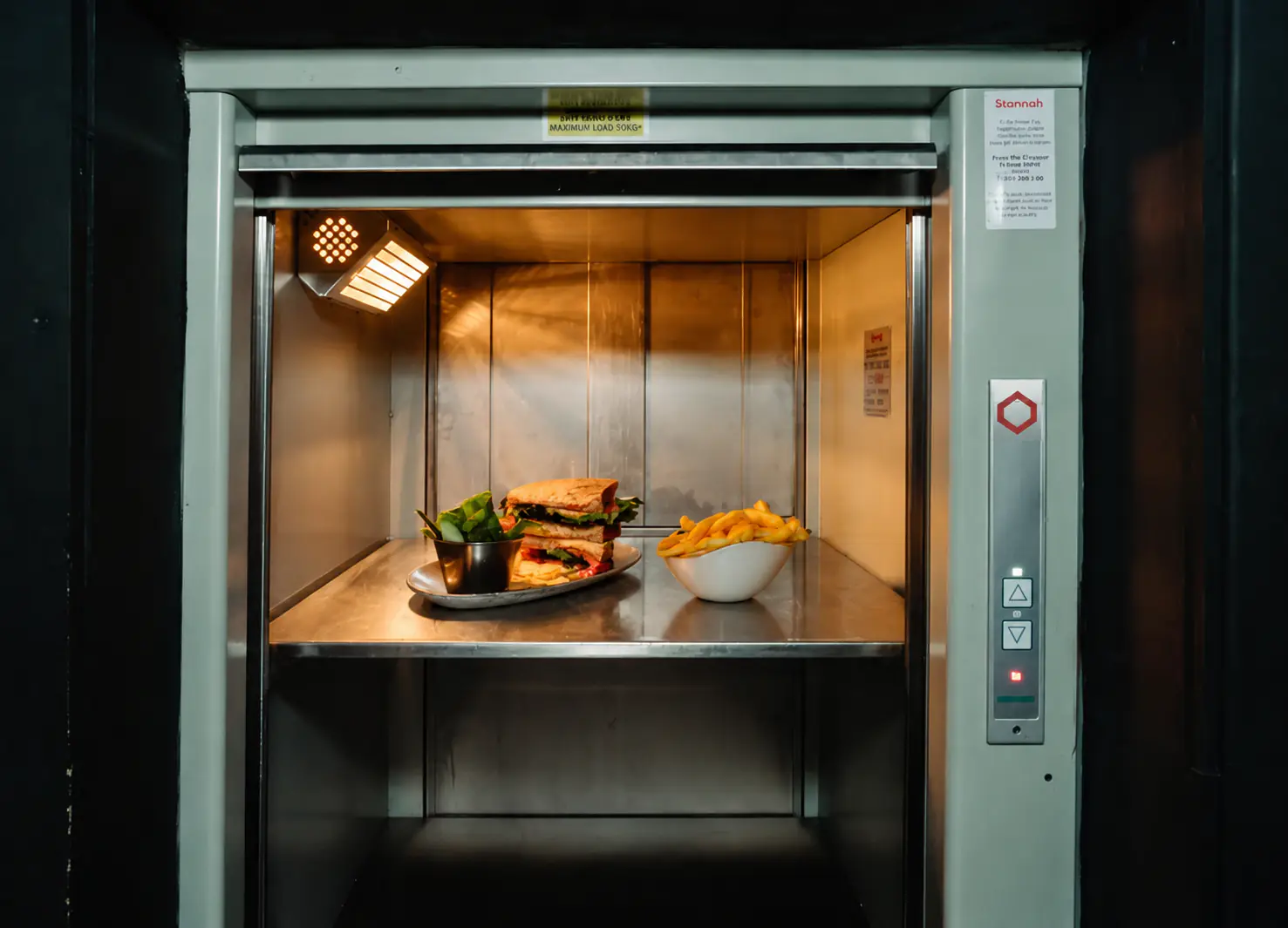 Interior of the stainless steel lift car showing a shelf holding a plated sandwich and a bowl of fries, with the control panel visible on the right wall.