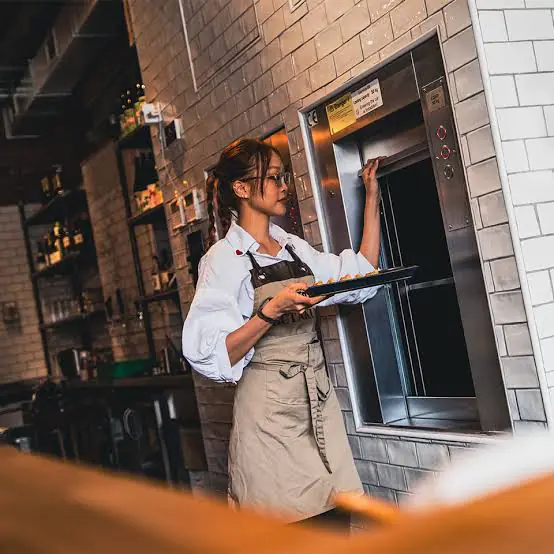A waitress in an apron loads a tray of pastries into a small stainless steel service lift set into a tiled kitchen wall.