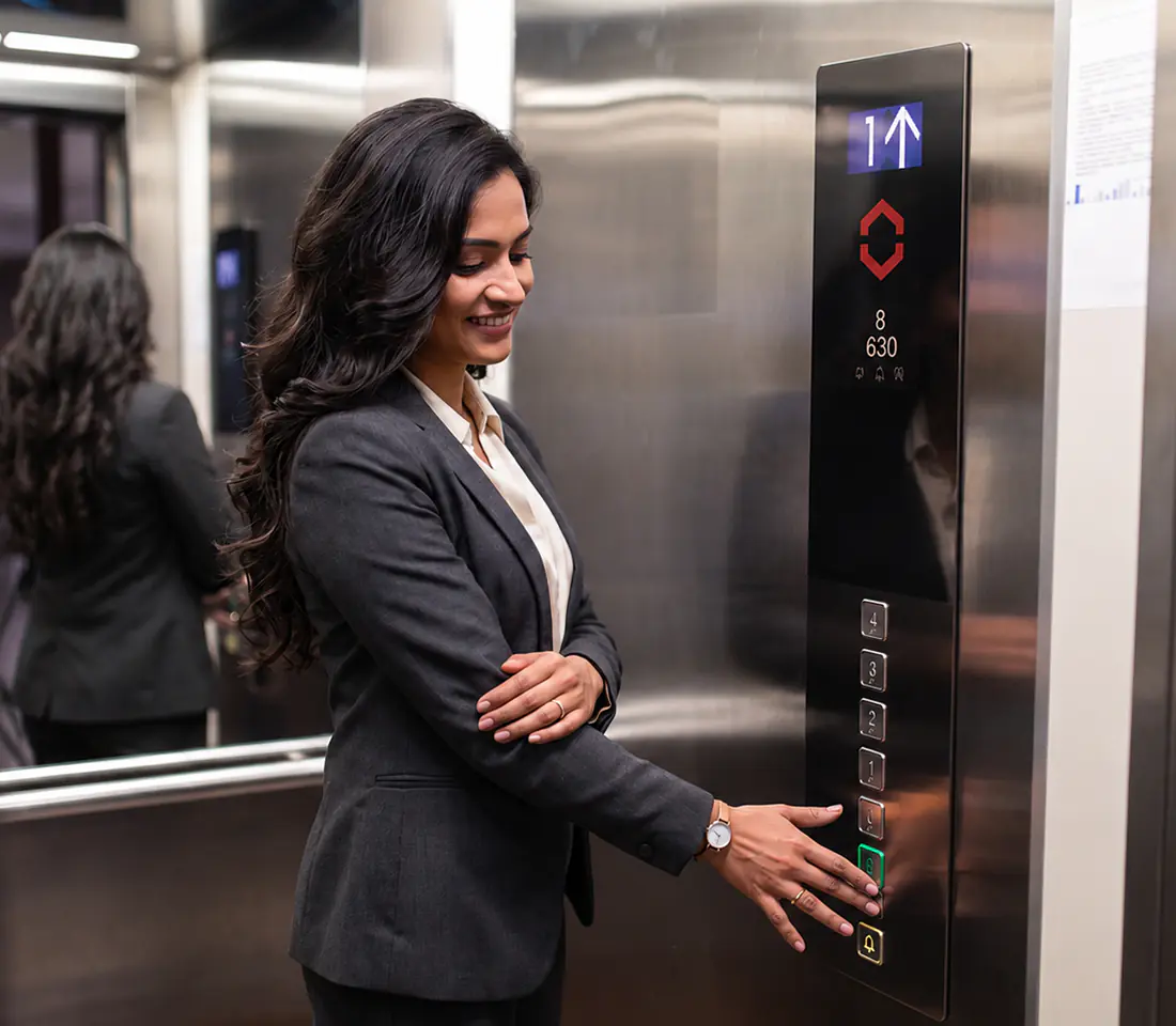 Businesswoman in a grey blazer pressing the floor call button on a stainless steel elevator control panel.