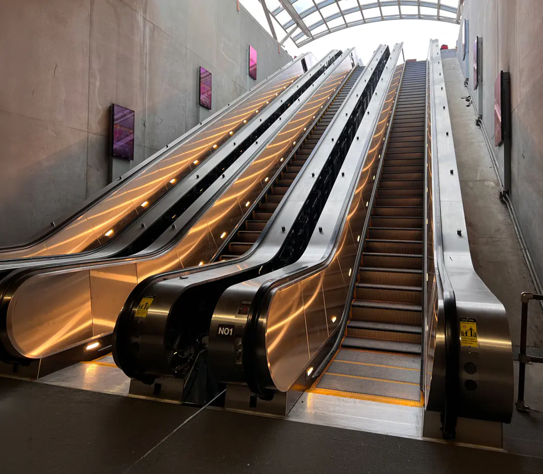 Frontal view of an empty stainless steel escalator with glass balustrades, shot from the base looking up into a bright glass walled interior.