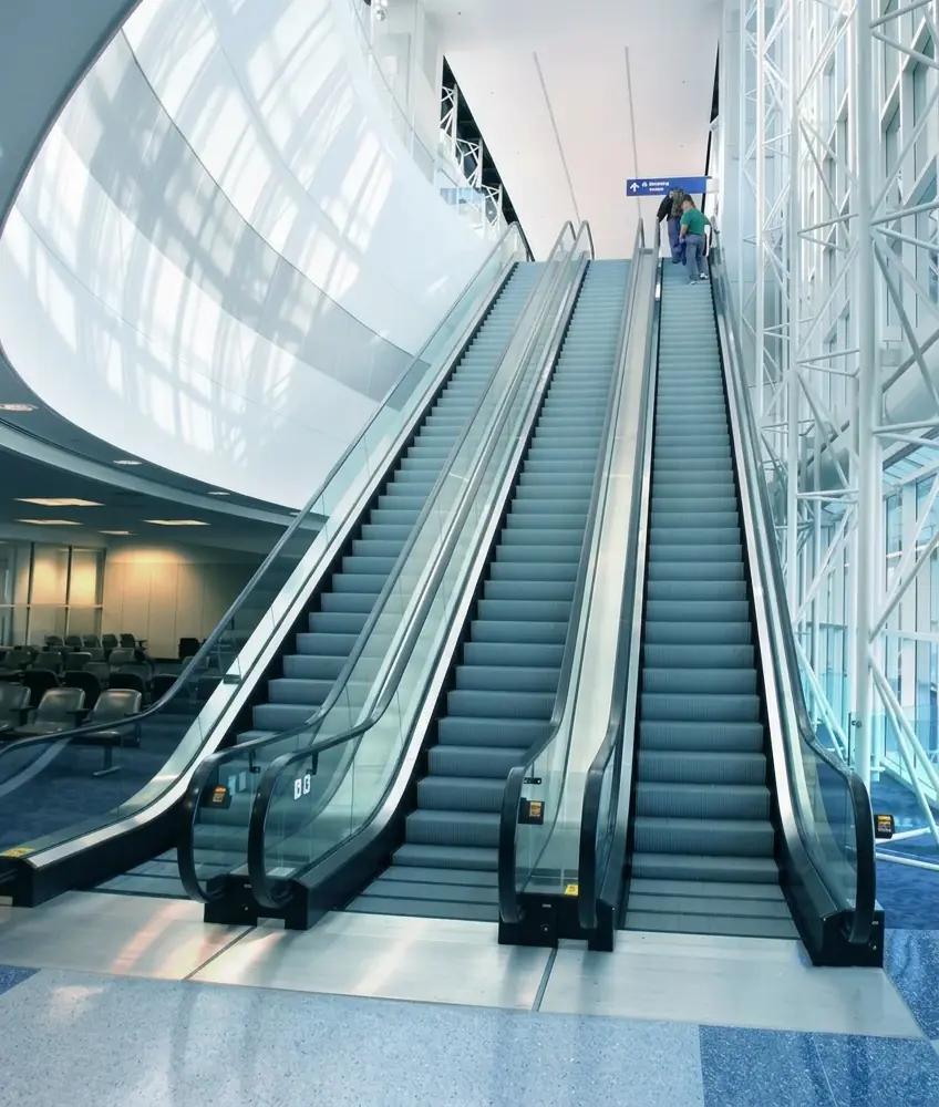 Wide shot of a three lane escalator bank in a large glass walled terminal atrium, with two people riding up near the top.