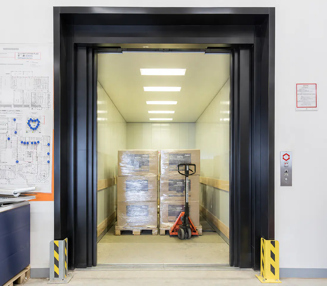 A freight lift with a dark steel frame and yellow and black corner guards, loaded with plastic wrapped pallets and a red hand pallet truck.
